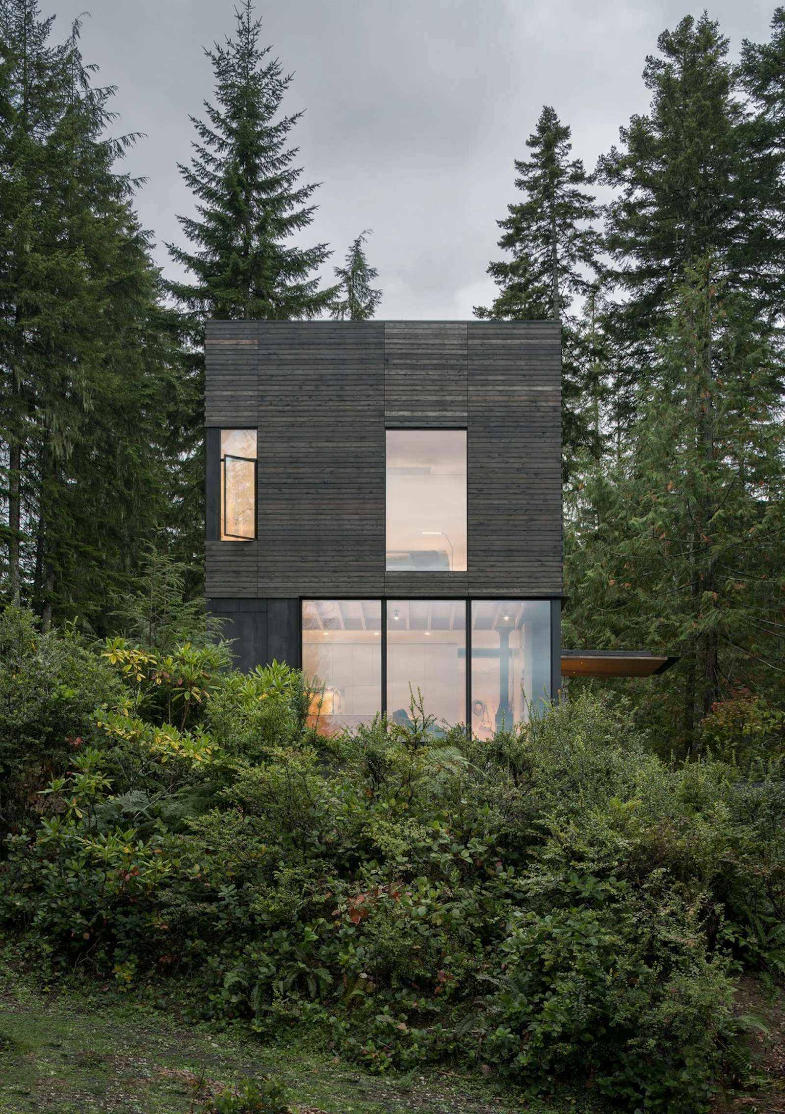 Petit chalet nordique minimaliste à toit plat de deux étages avec revêtements de lattes de bois horizontal couleur gris foncé, implanté dans une forêt verdoyante.