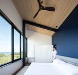 Bedroom, Dark Hardwood, Bed, Night Stands, Wardrobe, Storage, Ceiling, and Rug The master bedroom overlooks views of the water and beach through continuous glazing.  Bedroom Wardrobe Dark Hardwood Bed Photos from A Folded Steel Roof Shields a Fortress-Like Abode in Nova Scotia