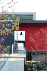 A small bridge connects the parking pad to the front entrance with a large pivot door. The home is primarily clad in Swiss Pearl exterior cement composite panels, Accoya timber, and painted metal panels on an open rain screen.