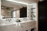 The master bathroom features a marble vanity with a mitered edge, with open shelves on the sides. To emphasize a minimalistic look, the custom steel-framed mirror is backlit with LED lights. A bi-parting sliding door made of black stained wood separates the toilet room and laundry room.