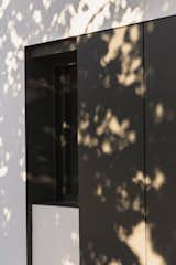 Detail of the façade where steel shutters integrate seamlessly into the white volume, enhancing the minimalist aesthetic. The interplay of natural light and shadows adds depth and movement to the architectural composition.