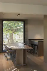 The dining area is framed by a large window, visually extending the space to the outdoors. Natural oak furniture and a suspended linear light by Santa&Cole enhance the warm and functional atmosphere.