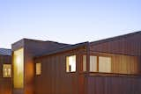 The top portion of a house at dusk. The house is covered in slatted cedar siding and the lights inside the house glow warmly.