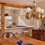 Dining room with custom table and built in marble and cherry wood dry bar looks into open kitchen separated by exposed beams. Kitchen boasts large countertop, open range, mix and matched bar stools, mosaic back splash, and ceiling high white cabinetry with gold hardware.