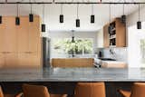 Macab quartzite slab island countertop looks into a kitchen with white oak wood flush cabinetry,  dark wood open shelving, white countertops and matching backsplash with matte black hardware and pendant lighting.