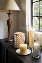 An antique lamp and votive candles rest on dark wood book shelves, against white paneled walls and shuttered windows in this living room.