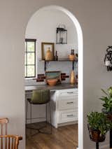 An arched doorway leads into the kitchen with cream and brown tiled backsplash, white cabinetry, black hardware, open shelving, and lantern sconces. The countertop also doubles as a desk with green leather bar stools.