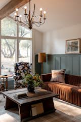 Cabin inspired living room with dark olive green wainscoting walls, a brown velvet couch, twin blue floral oversized chairs, plaid rug, a dark wood coffee table, and antique chandelier lighting.