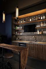 This moody game room boats a massive bar with dark blue walls, blue/grey backsplash tile, open shelving, dark walnut cabinetry, gold hardware and appliances, a built in mini fridge, frame tv, and its own bar counter with gold pendant lighting and leather stools.