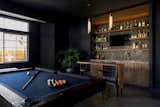 This moody game room boats a massive bar with dark blue walls, blue/grey backsplash tile, open shelving, dark walnut cabinetry, gold hardware and appliances, a built in mini fridge, frame tv, and its own bar counter with gold pendant lighting and leather stools. As well as a dark wood/blue felt pool table and drop down projector.