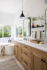 Ensuite bathroom with freestanding tub, medium-light wood cabinetry with black matte hardware and appliances, white counter tops, black matte metal twin mirrors and twin pendants.