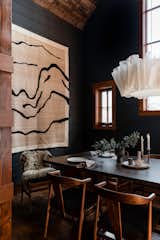 Farrow & Ball painted black paneled dining room with wrapped beams, paneled wood ceiling, a large dining table for eight, a mountain themed tapestry, and Anders pendant lighting.
