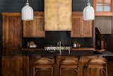 The mountain vibes are strong in this rustic industrial kitchen with textured dark wood cabinetry, an iron wrapped hood, milk globe pendants, black shiplap walls, and black counter tops and backsplash.