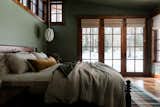 Ensuite bedroom with french doors gives out calming nature vibes through its green walls, midcentury furniture, layers of Parachute bedding, geometric pendant lighting, and multi colored plaid rug.