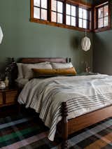 Ensuite bedroom gives out calming nature vibes through its green walls, midcentury furniture, layers of Parachute bedding, geometric pendant lighting, and multi colored plaid rug.