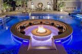 At the epicenter of the outdoor space is the circular fire lounge and patio that beckons guests from the moment they enter the home’s front door. Appearing to float within the pool, the 8 foot circular lowered fire lounge and patio area is constructed of tumbled travertine pavers in a French pattern. Cladded with glass tile and a two foot precast concrete cap, the glowing fire pit measure five foot in diameter. To enjoy the warmth and conversations, seating within the fire pit areas includes custom designed circular benches of tile with raised backing. Decorative pillows and cushions add to the comfort for viewing the entire patio area