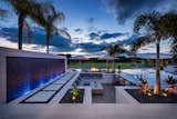 As sun sets, water glistens as it falls down the aria wall of oceanside tiles.  Incorporating a water feature. By Great American Waterfall Company.  The swim up bar lounge is bordered by stepping pavers and landscaping with FX Lighting. 