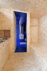 The OSB-clad living and sleeping area leads to a bold blue bathroom in this renovated home in Albino, in the Lombardy region of Northern Italy. The white fittings and fixtures in the bathroom echo the retro white cabinet handles used in the kitchen.