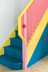 An angular yellow handrail folds over to meet a pink newel post, joined together by a red circular button. The stair is painted deep blue to accentuate its presence in the room. 