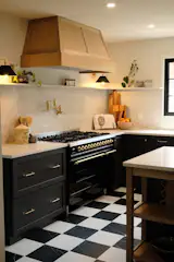 The brass details of the ILVE range are echoed in the brass hardware and tapware. Above the range, a timber-clad extractor offers a soft juxtaposition to the black cabinetry and marble splashback and countertops.