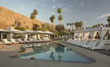 The central pool is surrounded by white loungers and private cabanas, with views of the nearby mountains and palm trees.