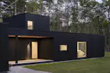 The composition of flat-roofed volumes—clad entirely in Nakamoto Forestry yakisugi sourced from PEFC-certified, air-dried Japanese cedar—reads as a single dark form against the forested landscape, punctuated by carefully placed openings that offer glimpses of the bright interior. At twilight, the entry facade (as seen here) becomes illuminated by warm light spilling from the covered porch and windows onto the gravel path.&nbsp;