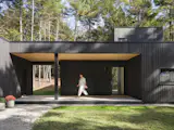 A generous covered entry porch provides a sheltered connection between the main house and a small garage and workshop. The warm-toned wood soffit creates a striking contrast against the charred Japanese cedar cladding, while the open breezeway beyond frames a view through to the woods.