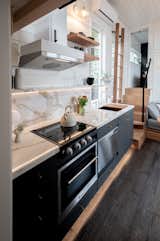 Gooseneck Tiny home with dark cabinets, accent lighting, brass hardware, white faucet, porcelain counters, and floating shelves by Tru Form Tiny