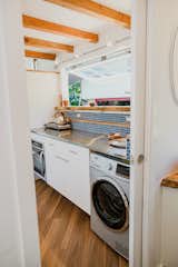 stainless steel appliances and stainless steel countertops in this galley kitchen design. Blue backsplash tile multi-level giving a whole other ledge to put spices on, and then you have the oversized think mango slab for a kitchen windowsill. Washer/dryer combo. Full-size fridge. Above the counter cook-top, built-in oven. stainless fixtures. Quartz composite sink. floating stainless kitchen shelves. Exposed stained ceiling beams supporting the loft.