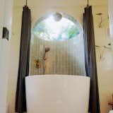 This luxury tiny home bathroom had a curved shower wall with a hand held and a rain shower head. sage tile and gold fixtures, this bathroom shower skylight is actually a dome to fit the curvature of the wall and round bathtub.