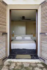 The practical bedroom is exquisitely minimal, with a built-in bed, hanging basket lamps, and a powerful overhead fan. The slate floor leads right out into the garden, the double doors opening to reveal a mountain of pine trees.