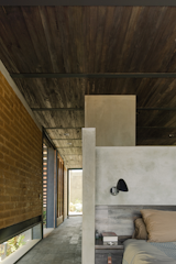The main bedroom with a bed, side table and lamp visible, as well as a long corridor running to the back wall of windows. A long horizontal window runs along the floor of the hallway. 