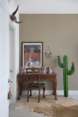 In Guta Louro’s office nook, a painting and books sit atop a wooden desk. Next to the desk is a metal cactus, hanging on the wall is a dreamcatcher, and on the floor is a cowhide rug. 