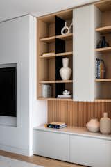 The living room features custom wall-to-wall millwork composed of light-toned cabinetry, natural oak elements, and black metal accents. A thoughtful interplay of open and closed volumes provides ample storage while visually anchoring one side of the room.