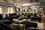 A generous, custom sectional draped in dark green and black sultry velvets, anchors the primary seating area. A black lacquer and brass coffee table from the Lori Morris collection and custom black velvet club chairs with brass accents, complete the vignette, gathered around a fireplace and oversized TV wall, clad in green back painted glass and brass strapping.