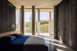 A bedroom has a window wall with a view of nature and a flat roof supported by rectangular columns.
