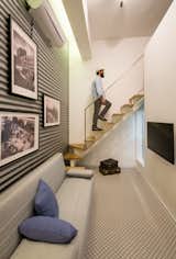 The photograph captures a person ascending a wooden staircase with a mesh railing within a modern interior space. The room is decorated with black and white framed photographs on corrugated metal walls, and there is a contemporary grey sofa with blue cushions and a vintage suitcase on the patterned floor. A flat-screen TV is mounted on the wall opposite the sofa, contributing to the room's urban chic aesthetic.