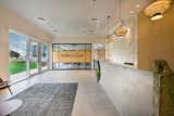 This image presents a wide-angle view of a modern, inviting lobby space. Natural light streams in from the large windows, complementing the lobby's neutral color scheme. The reception area features a textured concrete desk, wooden wall panels, and circular pendant lights that create a warm, welcoming atmosphere. The glass-walled conference room with the "Inspired to Cure" sign suggests a medical or scientific setting. The integration of indoor and outdoor space, along with the clean, contemporary lines, reflect a design that values transparency and a connection to nature.