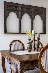 A large six-seater dining table commands attention, its carved details on the legs and side panels adding an air of grandeur and sophistication. Wooden chairs, upholstered in striped fabric, invite guests to linger and indulge in culinary delights, while a magnificent wooden Jharokha, intricately carved from mango teak, serves as a stunning focal point against the backdrop wall.