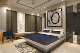 The room is elegantly decorated in a contemporary style. 
A large bed with a navy blue comforter and matching pillows takes center stage. The bed's headboard and footboard are a light beige or cream colour, appearing modern and minimalistic. 
The walls are a light beige tone and are paneled in vertical stripes. A framed abstract piece, predominantly black and white, hangs on the wall above the bed. 
Large, taupe-coloured curtains cover the windows and allow some natural light to filter in. 
A light beige or cream-coloured rug is laid on the floor. The flooring throughout the room appears to be from the same light beige/cream colour family, likely a type of polished stone or tile.
A dark-blue upholstered armchair is placed near the door, and a light beige/gold-toned bench is situated beside the window.
A portion of a dark grey or black cabinet/wardrobe is visible to the right of the bed.
The ceiling has a decorative molding with recessed lighting. Also, a ceiling fan is present. The lighting in the room seems well-distributed and balanced.
The overall impression is of a spacious and stylish bedroom, potentially part of a hotel or a luxurious residence.