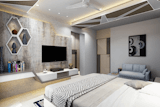 The wall behind the television unit features a textured, mottled greyish-tan surface resembling a decorative plaster or wallpaper. Three white hexagonal wall shelves, each containing a decorative vase, are mounted on this wall. 
A modern, flat-screen television is mounted on the wall above a sleek white and grey entertainment center/TV stand. The stand has a segmented design with a slightly angled top section. A stack of books rests on the top of a white section of the stand. 
The bedroom is furnished with a bed that takes up most of the width of the room, covered with a light-grey bedspread with a geometric, zig-zag pattern.  A light-grey/blue couch is placed near the foot of the bed. A framed abstract artwork piece, displaying black and white circles and shapes, hangs on the wall above the couch. 
The ceiling has a contemporary design with light grey panels creating a geometric pattern and recessed lighting is visible. A ceiling fan is also present. 
The overall colour scheme is muted and sophisticated, using tones of grey, white, and beige. The room has a clean and minimalist aesthetic.