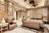 The room features off-white/beige-toned walls, with a textured pattern that appears to be a type of mosaic or tile.  The ceiling has a similar light beige tone with a sculpted, wave-like pattern.  A ceiling fan with wooden blades is centrally located. A projector is visible within the ceiling fan's assembly.
A large upholstered bed with a light beige/cream headboard, tufted and ornate, takes center stage.  The bed has a matching footboard and appears to be very sizeable.  Purple accent pillows are on the bed.  A matching bedside table is present on the right side of the bed.
A light beige/cream coloured armchair, with a slightly sculpted back and armrests, sits to the left of the bed area. A round, light beige/cream-coloured  table is positioned in front of it, with a vase holding a small bouquet of yellow/gold orchid-like flowers.
Framing the room are two large framed prints featuring dried floral designs in cream tones hanging on the walls.
A marble or polished stone floor covers the entire room. A patterned rug is placed under the bed, in a dark brown and off-white floral design.  A small section of a vanity or desk area is visible near the back wall. 
The overall style is sophisticated and elegant, with a calm colour palette and detailed furnishings.