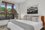 A serene bedroom featuring a king-sized bed dressed in crisp, monochromatic linens. The room offers seamless access to a lush outdoor terrace and panoramic views of the surrounding forest, with warm wooden-framed windows enhancing the connection to nature. A textured wall hanging adds a boho-chic aesthetic, complementing the minimalist design.