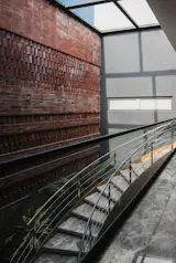 Angled perspective of a stairway under a skylight, flanked by textured red bricks and smooth gray surfaces.