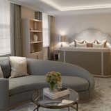 This beautifully designed turnkey suite exemplifies modern elegance and comfort. Featuring a soft, neutral palette, the space seamlessly blends a curved upholstered sofa, a sleek glass coffee table, and a luxuriously dressed bed with an upholstered headboard. Carefully curated decor, including books, greenery, and artistic shelving accents, enhances the refined ambiance. Natural light filters through double-layered curtains, adding a touch of softness to the room. A perfect example of how a complete turnkey approach can deliver stylish, cohesive results efficiently and cost-effectively