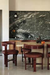 Dining-rich wooden chairs encircle a sleek stone table, set against a dramatic green marble backdrop, blending luxury with organic warmth.