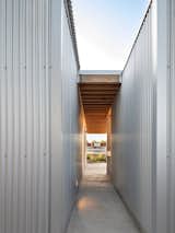 A open air hallway between two corrugated volumes creates a framed view toward the street and garden. The exposed wood roof structure and minimal detailing highlight the building’s straightforward construction approach.