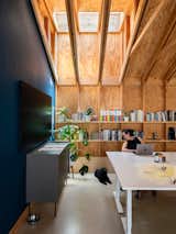 A skylight draws daylight deep into the workspace, illuminating the raw wood structure and built-in shelving. The casual presence of plants, books, and even the studio dog reflects the building’s approachable scale and lived-in comfort.