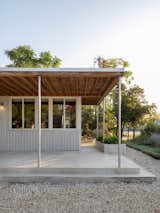The porch’s exposed wood ceiling and galvanized fence-post columns embody the project’s ethos of using straightforward, readily available materials. The planter at the edge frames the entry and softens the transition from gravel drive to interior.