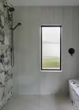 An expanded shower features large-format tile and dramatic stone detailing. A newly added reeded-glass window filters natural light into the space, highlighting the contrast of textures and matte black fixtures.